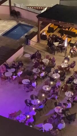 The video shows an aerial view of an outdoor seating area at night. There are numerous small round tables with chairs around them, occupied by many people. Most of the people appear to be adults, with a mix of genders. Some tables have drinks or small light fixtures on them. There is a pool to the left of the seating area. A covered area with lounge seating is visible in the background, also with people seated. String lights and purple lighting are illuminating the scene. The overall impression is of a busy, social outdoor establishment.