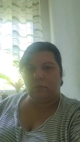 A woman with dark hair is looking directly at the camera. She is wearing a grey and white striped t-shirt. The background shows a window with white curtains and a plant.