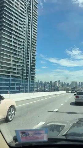 A vertical, first-person perspective video shows a view from inside a vehicle traveling on a multi-lane highway. The highway has a white barrier on the left and a concrete barrier with a reflective fence on the right. A tall, modern building with many balconies is visible on the left. In the distance, a cityscape with numerous tall buildings is seen under a bright blue sky with scattered white clouds. Several cars and a white van are on the highway, moving in the same direction as the camera. The front dashboard of the vehicle is visible at the bottom of the frame, with a sticker in the lower left corner. The weather appears to be clear and sunny.