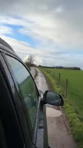 From the perspective of someone inside a moving, possibly black, car, the video shows a rainy day scene as the vehicle travels down a narrow, muddy, unpaved road. A field of bright green grass runs alongside the road, bordered by a wire fence. The cloudy sky is visible, with some blue patches. The car's wet window and side mirror are prominent in the foreground. A bare tree is visible in the distance.