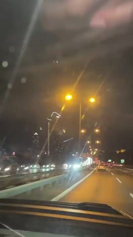 The video shows a nighttime view from the perspective of a driver in a vehicle, looking out through the windshield. The vehicle is on a highway with multiple lanes of traffic. Streetlights illuminate the road, casting a warm yellow glow. On the left side of the highway, there are buildings, including a tall skyscraper adorned with lights. The sky is dark, with some clouds visible. The headlights and taillights of other vehicles create streaks of light as they move along the road. The scene suggests a city at night, with the highway being a major artery.