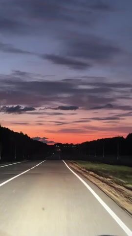 The video shows a POV shot from a vehicle driving on a road at dusk. The sky is filled with a colorful sunset, with hues of orange, pink, and purple. The road ahead is illuminated by headlights, and there are trees lining both sides of the road. The scene is calm and peaceful, with no people or other vehicles visible.
