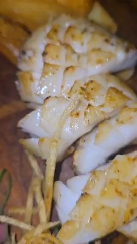 A close-up shot captures a delicious meal served on a wooden platter, featuring grilled fish cut into pieces, with some small fries on the side, a white-colored crispy snack, and a dark dipping sauce in a small bowl. The platter is placed on a wooden table, with other plates visible around it, indicating a dining setting.