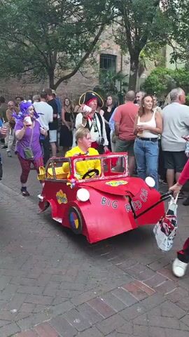 A group of people are walking down a cobblestone street, possibly in a parade or festival. In the foreground, a person in a blue shirt and black pants walks alongside a red toy car. The car has "BIG RED CAR" written on it and a person in a yellow shirt is sitting inside. Behind them, a person in a purple shirt and plaid pants is holding a drink and wearing a purple wig. To their right, a person dressed as a pirate with a hat and vest is visible. Further back, a person in a green dinosaur costume is walking. Spectators are lining the street, watching the procession. The environment appears to be an urban setting with trees and buildings visible in the background.