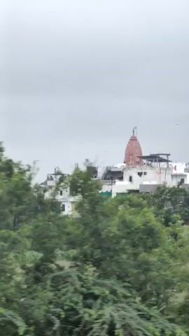 The video shows a blurred view of a Hindu temple with a pink spire. The temple is situated on a hill and surrounded by green trees. The sky is overcast and grey. The camera appears to be moving, causing the motion blur in the footage. The buildings around the temple are also visible, but are similarly blurred.