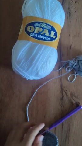 The media file is a close-up shot of hands crocheting with gray yarn on a wooden surface. A large ball of white yarn with an "OPAL Dört Mevsim" label is visible to the left. A smaller ball of gray yarn is in the background. The lighting suggests it is daytime. The hands are actively engaged in the crocheting process, with the crochet hook moving through the yarn to create stitches. The focus is on the hands and the craft, implying a moment of creation or hobby. The setting appears to be indoors, given the presence of yarn and the indoor lighting. The "Dört Mevsim" text on the yarn label translates to "Four Seasons" in Turkish, indicating the yarn is suitable for use year-round. The location context of Batken, Kyrgyzstan, does not provide any visual cues within the media file itself.