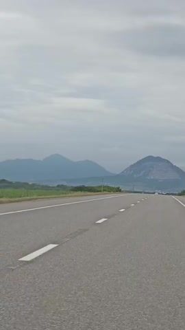 A point-of-view shot shows a paved road with white dashed lines. Mountains are visible in the distance under a cloudy sky. The road curves slightly to the right. Cars are driving on the road in the distance. A single traffic sign is visible on the left side of the road.