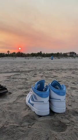 The video shows a pair of white and blue high-top sneakers resting on a sandy beach. The sun is setting in the background, casting an orange glow across the sky. The scene is tranquil and peaceful, with the gentle waves of the ocean likely in the distance. The sneakers appear to be new or well-maintained.
