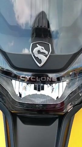 The video shows a motorcycle, likely a Cyclone model, which is blue and yellow, equipped with off-road tires and a clear windscreen. The camera focuses on the front of the motorcycle, highlighting the headlight, logo, and branding. Later, it zooms out, revealing more of the vehicle's design and accessories such as sidebars and a mounted storage box. The motorcycle is parked outdoors on pavement, and the background includes parked vehicles and a woman standing nearby.