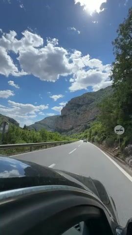 The video shows a car driving on a two-lane road through a mountainous area under a bright, partly cloudy sky. The road is paved and has white lane markings. A metal guardrail runs along the left side of the road. The foreground includes the hood of the car from which the video is being taken. In the distance, another car is visible driving in the same direction. The mountains in the background are rocky and covered with green vegetation. The sky is blue with scattered white clouds. The sun is shining brightly, creating a glare. Trees line the roadside.
