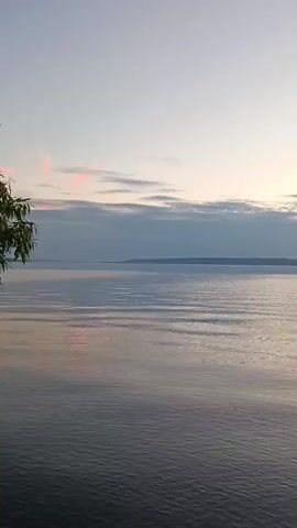The video displays a wide shot of a large body of water, likely a lake or ocean, under a sky that appears to be at either sunrise or sunset. The water's surface exhibits gentle ripples, suggesting a calm or lightly breezy condition. In the distance, a low-lying landmass or horizon line is visible, partially obscured by atmospheric haze. The sky transitions from a pale, almost white hue at the top to softer tones of blue, gray, and hints of pink and orange closer to the horizon. Clouds are present, appearing as elongated, soft bands that run horizontally across the scene. There are no people, boats, or man-made structures immediately apparent in the frame. The overall impression is one of natural tranquility and expansive natural beauty.