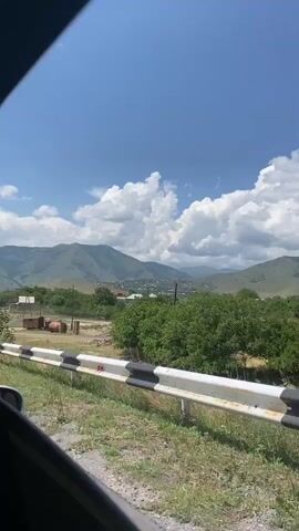 The video shows a view from inside a vehicle, looking out of a window. The vehicle is moving, as indicated by the blurred foreground and the shifting scenery. In the distance, there are mountains under a blue sky with white clouds. In the mid-ground, there are trees and some buildings, suggesting a rural or semi-rural area. A guardrail with black and white stripes is visible in the foreground, running along the side of the road. The vegetation appears green and lush, indicating a warm season. There are no people visible in the video.