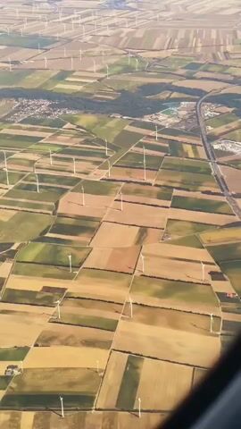 This is an aerial video of a vast agricultural landscape dotted with numerous wind turbines. The land is divided into various rectangular fields, some appearing green and others a golden-brown, suggesting different crops or stages of growth. A highway runs through the scene, and in the distance, a cluster of buildings can be seen. The wind turbines, which are white with three blades, are spread across the fields, some in groups and others individually. The video is taken from a high altitude, likely from an airplane, as indicated by the slight angle and the presence of a portion of an aircraft wing in the bottom left corner of the frame. The lighting suggests it is daytime, possibly late afternoon due to the warm tones of the landscape.