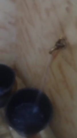 The video shows water flowing from a brass faucet into two black plastic buckets placed on a tiled floor. The walls are covered with light brown and white marble-like tiles. The faucet appears to be old and slightly tarnished. The water stream is steady, filling one of the buckets noticeably more than the other. The scene is shot in a static, close-up view.