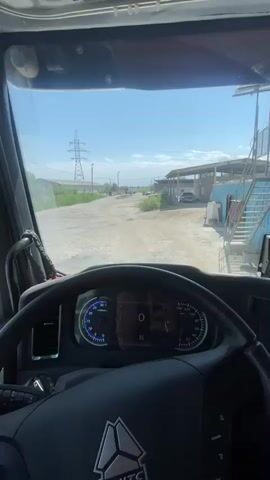 The video shows the interior of a truck cab, looking forward through the windshield. The driver's side mirror is visible on the left, reflecting a red truck and a building. The steering wheel and dashboard are in the foreground. Outside, a dusty road leads into an industrial-looking area with buildings and a power line tower. A person is sitting on a set of stairs on the right side of the frame.
