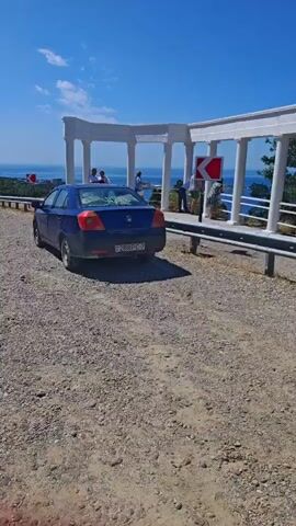 A blue car is parked on a gravel shoulder next to a paved road. The car is facing away from the camera. In the background, there are people standing near a white structure with columns, possibly a gazebo or pavilion. Beyond that, a body of water, likely the sea, is visible under a clear blue sky. There are guardrails along the side of the road, and traffic signs indicating sharp turns are also present. The terrain appears to be hilly, with vegetation on the left side of the road. Another car is seen driving away on the road in the distance.