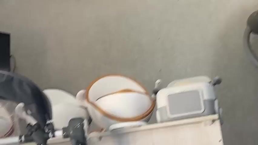 A short, blurry video shows a shelf with various objects. On the left, part of a stroller or baby carrier is visible. In the center, a white and orange plastic basin or bowl is prominent. To the right, a gray and white electronic device, possibly a radio or speaker, sits on the shelf. The background is a textured, light-colored wall. The lighting is diffused, and the focus is unclear, making precise identification of some items difficult.