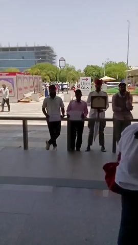 The video shows an outdoor scene that appears to be a transportation hub, possibly an airport or a train station. In the initial shot, four men are standing behind a railing, each holding something. One man is wearing a traditional hat and holding a framed certificate. The others are holding papers. In the background, there is a sign for "Uber OLA". The scene then transitions to show a wide corridor with people walking by, some with luggage. There are seats along the corridor where people are waiting. The area is covered with a roof supported by pillars and beams. There is an advertisement on the roof that says, "Land your brand in the spotlight." Overall, the video seems to depict a busy transportation area with people arriving and departing.