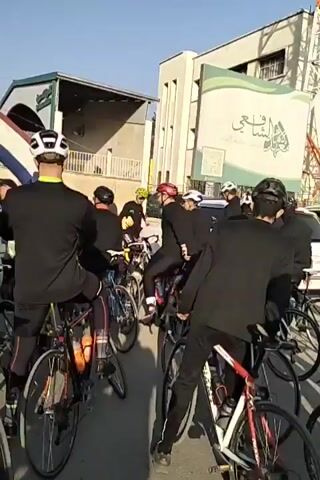 A crowd of people, many on bicycles and wearing helmets, are gathered on a street under a blue sky. An arch in the colors of the Dutch flag stands at the front of the crowd. Most people are wearing black sweatshirts. Some people, including a young boy with a green baseball cap, turn to look at the camera. A man with glasses and a helmet gives a direct stare into the camera. Buildings are visible in the background.