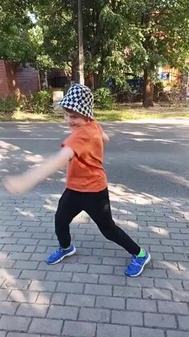 A young boy wearing an orange t-shirt, black pants, a checkered bucket hat, and blue sneakers is performing martial arts moves on a brick sidewalk next to a paved road. He is in a tree-lined area with buildings in the background. The boy is captured in various dynamic poses, including kicking and punching. The video appears to be a short clip documenting his martial arts practice.