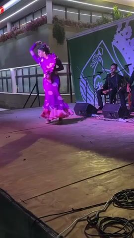 A flamenco dancer performs on a wooden stage at night, wearing a vibrant purple dress with pink polka dots and ruffles. Her dress flares out as she moves, and she has her hair styled up with a floral accessory. She is positioned on the left side of the stage, with her arms raised and her body angled away from the camera. Behind her, a green banner with a white logo is visible. To the right of the dancer, two men are seated on the stage. One man, wearing a black shirt, is playing a guitar. Another man, also in black, is seated next to him, possibly singing or playing another instrument. Stage lighting illuminates the scene with pink and purple hues, casting shadows on the wooden floor. Cables are coiled on the lower right corner of the stage. In the background, a building with illuminated windows and a balcony adorned with plants can be seen.