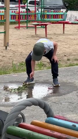 A boy wearing a light blue t-shirt, dark pants, a dark green baseball cap, and athletic shoes is on a paved surface near a sandy area. He is bending over and using a twig to gather a pile of green leaves and debris. The boy is focused on his task and appears to be creating a small pile of leaves. In the background, there is a playground with colorful structures, benches, and cars. The overall environment suggests an outdoor public space like a park or playground.