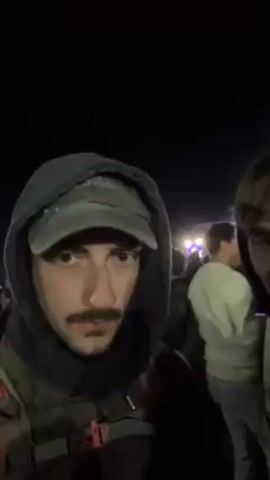 Two young men are recording a video selfie at an outdoor music festival at night. They are surrounded by a crowd of people, and a stage with bright lights is visible in the background. One of the men is wearing a hooded sweatshirt and a baseball cap, and the other is wearing a hooded sweatshirt. The man in the cap briefly gives the middle finger to the camera. The crowd is moving and swaying to the music.