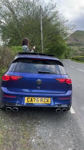 A woman is standing out of the sunroof of a blue Volkswagen Golf R, holding her phone up and taking a picture or video. The car is parked on the shoulder of a two-lane road. The background shows a green and rocky mountain landscape under a cloudy sky. A wooden fence separates the road from the greenery, and a small drainage grate is visible next to the fence.