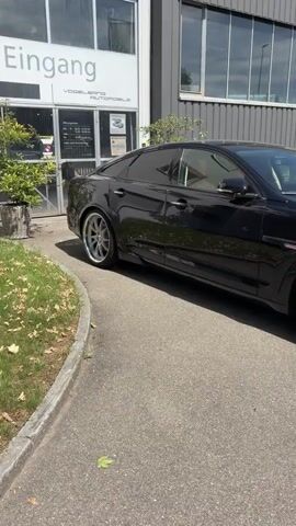 The video shows a black Jaguar XJ parked outside a building. The car is seen from the side, with a focus on its sleek design and custom wheels. The building appears to be a business or workshop, with large windows and a grey facade. The environment is outdoors, with pavement and some greenery visible. The camera pans across the car, highlighting its features. There are no people or any other significant objects or actions to note. The video appears to be a showcase of the car.
