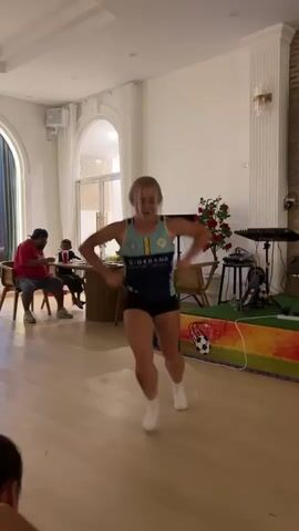 A woman in athletic wear is dancing in a room. She starts by squatting and then moves to a crawling position on the floor before rising back to a squat. She has blonde hair tied back and is wearing a blue and yellow athletic top and black shorts. In the background, two men and a child are seated at a table. There is a keyboard and speaker setup to the right of the dancer, and a colorful banner with a soccer ball on it. The room appears to be a dining or event space with wooden floors and white walls.