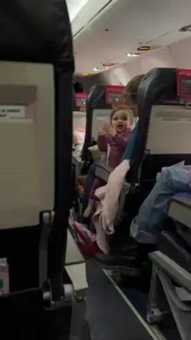 The video shows a child sitting on someone's lap on an airplane. The child is wearing a purple top and dark pants, and a pink blanket is draped over the seat. The person holding the child is wearing a teal top. Another person in the next seat is wearing jeans. The seats are black and gray. There are safety information cards and magazines in the seat pockets. The airplane cabin appears to be brightly lit. The child is clapping and moving their arms.