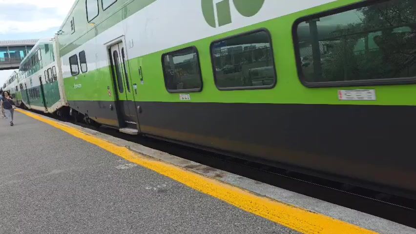 A GO train with green and white double-decker cars is stopped at a station platform. The platform has a yellow safety line. A few people are visible on the platform. One person is walking towards the train. Another person is walking away from the train. The train cars have multiple windows and doors. The train cars have "GO" branding on them.