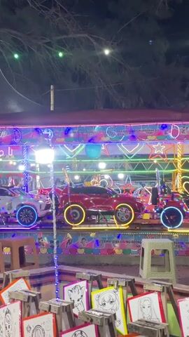 The video shows a night scene at an outdoor amusement or fairground attraction. The main focus is a row of colorful, toy-like cars, possibly part of a carousel or a small amusement ride. These cars are brightly illuminated with neon lights, featuring red, blue, and yellow accents, particularly around their wheels and bodies. They are positioned on a raised platform that is also decorated with colorful lights and festive designs. In the foreground, there are several small wooden stands displaying coloring pages, some featuring cartoon characters. The overall ambiance suggests a lively and festive nighttime event, possibly catering to children. The background is dark, with hints of trees and outdoor lighting.