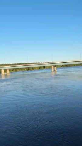 The video shows a wide river with calm blue water under a clear blue sky. A long, modern bridge spans across the river, supported by concrete pillars. On the far bank of the river, there is a line of trees. On the near bank, on the right side of the frame, a few trees are visible. The scene appears to be a natural landscape with man-made infrastructure. No people are visible in the video. The overall impression is peaceful and serene.