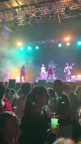 A band performs on a stage in front of a crowd. The band consists of five women. One woman is playing a saxophone. Two women are singing and dancing. One woman is playing a guitar. One woman is playing drums. The stage is lit with purple and green lights. The crowd is dark and blurry, with some people visible in the foreground.