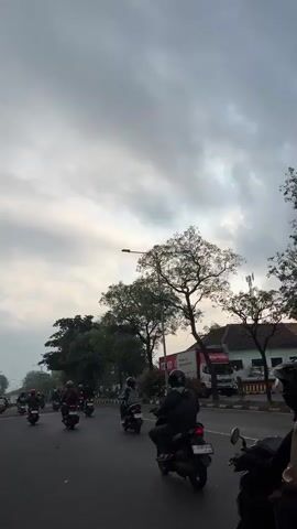 The video shows a busy street scene with multiple motorcycles and riders moving in the same direction. The sky is overcast with dramatic clouds. Trees line the side of the road, and in the background, there are buildings and what appears to be a truck. The road is paved and appears to be a public thoroughfare. The lighting suggests it could be early morning or late afternoon. The focus is on the movement of the motorcycles and the atmospheric conditions.
