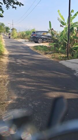 This media file captures a scenic view of a rural road in Kwaren, Indonesia. The scene is dominated by an asphalt road stretching into the distance, flanked by lush green grass and dry straw on the left, and dense vegetation, including banana trees, on the right. A black SUV is parked on the side of the road, partially obscured by the foliage.

A single individual on a motorcycle is traveling down the road, casting a distinct shadow. The time of day appears to be morning or late afternoon, indicated by the long shadows cast across the road and the bright, clear sky. The weather seems pleasant, with a clear blue sky and ample sunlight.

The overall impression is one of tranquility and everyday life in a rural Indonesian setting. There are no visible signs or text in the media file. The focus is on the natural landscape, the road, and the solitary motorcycle rider, creating a sense of peaceful journey.