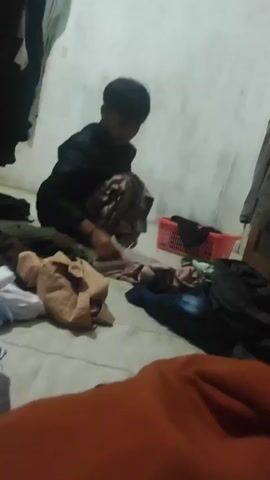 The video shows a young person squatting on the floor, sorting through a pile of clothes. The environment appears to be a room, possibly a bedroom, with clothes strewn around. There is a laundry basket in the background. The person is wearing dark clothing and is focused on the task of folding or organizing the garments. The lighting is dim, suggesting it might be nighttime or the room is not well-lit. The person picks up a piece of clothing, inspects it, and then places it back down or folds it. The action is slow and deliberate.