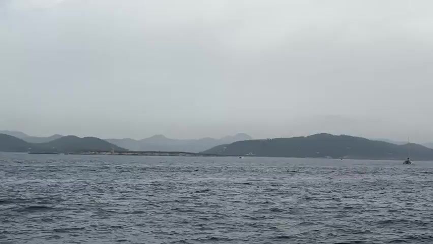 The video shows a wide view of the sea with distant, hazy mountains under a cloudy sky. The water is choppy with visible waves. Several boats are scattered across the sea, and on the distant shoreline, buildings and trees are visible. The overall atmosphere is muted due to the overcast conditions.