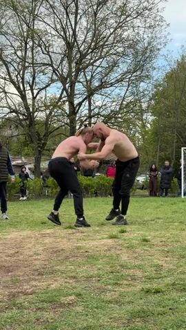 Two shirtless men wearing black pants wrestle in a grassy clearing. They grapple each other, arms intertwined, maneuvering for position. A small crowd watches from the perimeter of the clearing. Bare trees stand behind the wrestlers, and buildings are visible in the background. The scene is set outdoors, in daylight.