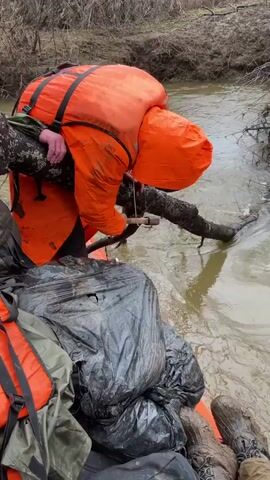 A person wearing an orange rain suit and life vest is perched on a raft in a muddy river, using a small axe to chop a tree branch that is lying across the water. Bags covered in black plastic and additional life vests are visible on the raft. The river is flowing and the surrounding area is wooded.