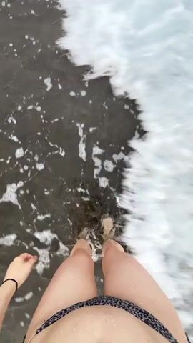 A first-person perspective video shows a person's legs and lower torso from the waist down, standing in shallow ocean water. The person is wearing a leopard-print bikini. The water is dark and choppy with white foam from waves washing over their feet and ankles. The camera is pointed down at an angle, capturing the movement of the water and the person's legs. The person's left hand is visible on the left side of the frame, resting on their upper thigh.
