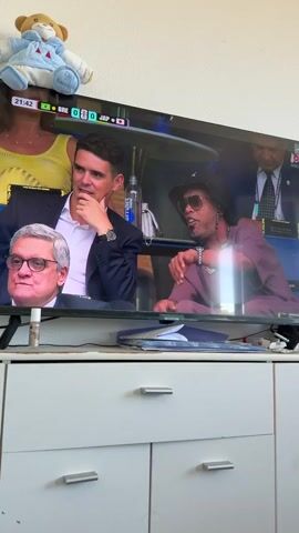 The video shows a television screen displaying a live broadcast of a football match. The score indicates Brazil (BRE) 0 - 0 Japan (JAP), with the time at 21:45. Three individuals are visible on the screen. The person on the far left is a man with grey hair, wearing glasses. To his right is a man in a dark suit, resting his chin on his hand, appearing thoughtful. On the far right is a person wearing a hat and sunglasses, dressed in a purple top and a pearl necklace, gesturing with their right hand. In the background of the broadcast, other people and the stadium setting are visible. A plush teddy bear toy is positioned on top of the television. Below the television, a white dresser with silver handles is visible, suggesting the setting is a home environment. The lighting appears to be indoor, and no weather conditions are discernible. The overall mood on the screen seems focused on the match and the commentators or guests discussing it. Visible text includes the score, time, and team abbreviations.