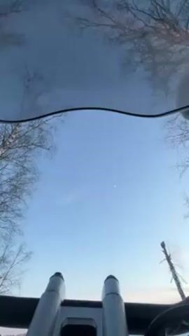 The video, shot from a low angle inside a vehicle, shows a person waving at the camera through the sunroof. The person is wearing a light beige hooded jacket, possibly with fur trim, and what appears to be a black face covering or mask. They have a backpack on. Outside, there are bare trees against a blue sky, with a small crescent moon visible. The vehicle interior shows part of the steering wheel and two cylindrical objects. The overall environment looks like a cold, possibly winter setting.