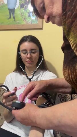 A woman sits on a couch while another woman helps her take her blood pressure. The sitting woman wears a white t-shirt with pink letters, glasses, and a stethoscope around her neck. She holds a black blood pressure monitor in her left hand. The second woman, with short red hair and a brown and green patterned shirt, is putting the blood pressure cuff on the sitting woman's left arm. A framed painting of a man and two women toasting with wine hangs on the wall behind the couch.