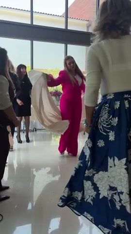 The media file shows a group of women dancing indoors. The foreground is dominated by a woman in a white long-sleeved shirt and a flowing blue skirt with a white floral pattern. Her back is to the camera as she twirls. To her left and in the background, other women are visible. One woman, wearing a black dress and black stockings, stands and watches. Another woman in a bright pink pantsuit with a matching jacket is dancing enthusiastically. A fourth woman, wearing a light yellow top and a patterned dress, is also visible to the right.

The setting appears to be a well-lit indoor space with large windows offering a view of greenery and a tiled roof structure outside, suggesting a daytime event. The flooring is reflective, creating mirror-like surfaces. The overall atmosphere seems festive and joyful, with the women engaged in energetic movement. The presence of tables with white tablecloths and floral arrangements in the background hints at a celebratory occasion, possibly a party or gathering. The location context is Santiago Yancuitlalpan, Mexico. No visible text is present in the media.