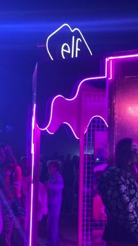 The video shows an Elf Cosmetics branded makeup station. The station is bathed in bright pink and purple neon lights, with the brand’s logo prominently displayed above the entrance. Inside the station, a makeup artist is applying makeup to a female customer. The customer is wearing a black skirt and a white patterned jacket. The makeup artist is wearing a black jacket with the Elf logo on the chest. The station is well-lit with a mirror adorned with round lightbulbs. Various makeup products are arranged on the table. The backdrop features the words "EXPRESS YOUR E.L.F." in large letters, with beaded pink curtains. Outside the station, a crowd of people can be seen, indicating a busy event or festival setting.