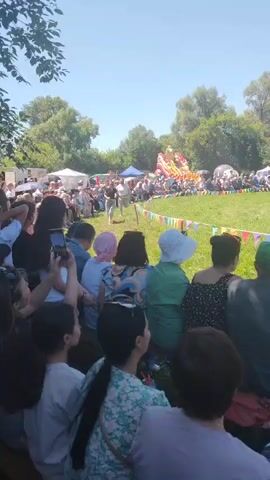 The video shows an outdoor event with many people gathered on a grassy field. A group of people are in the center of the field, some appearing to be performing or participating in an activity. There are tents and trees in the background, suggesting a park or fairground setting. A colorful banner with flags is strung across the middle ground. Spectators are seated on the ground in the foreground, some watching the event and others holding up phones to record. The sun is shining, indicating a clear day.