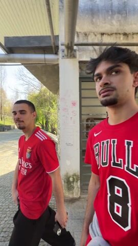 The video shows two men and a woman walking on a cobblestone pathway, presumably in a park or recreational area, on a sunny day. The first man is wearing a red Benfica soccer jersey with black pants, and is holding a phone. He looks briefly at the camera. The second man is wearing a blue and red shirt, grey sweatpants, and a black hoodie. The woman is wearing a red shirt and jeans. The walkway is partially covered by a metal and concrete structure, with stone walls on either side. There are trees and a road visible in the background. The camera follows the men as they walk along the path.