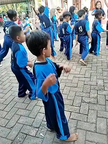 The video shows a group of young children, dressed in matching blue and black athletic uniforms, participating in an outdoor activity. The children are standing on a brick-paved surface. The child in the foreground, facing away from the camera, is wearing a blue shirt with black sleeves and a black number on the back. They are wearing dark blue pants with a light blue stripe down the side. The other children in the background are also wearing similar uniforms. Some of the children appear to be engaged in a coordinated movement, possibly a dance or exercise routine. The environment appears to be a schoolyard or an open area with some greenery and buildings in the background. The lighting suggests it is daytime.